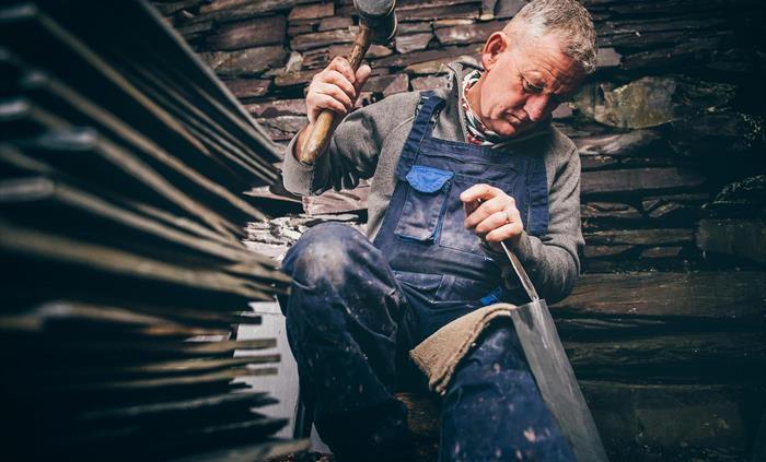 A man in a grey jumper and blue dungarees, sits beside a pile of slates. His right hand is raised, holding a wooden mallet and his left hand holds a c